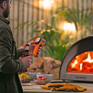 A man is holding the digital thermometer and getting a temperature reading from a lit igneus pizza oven