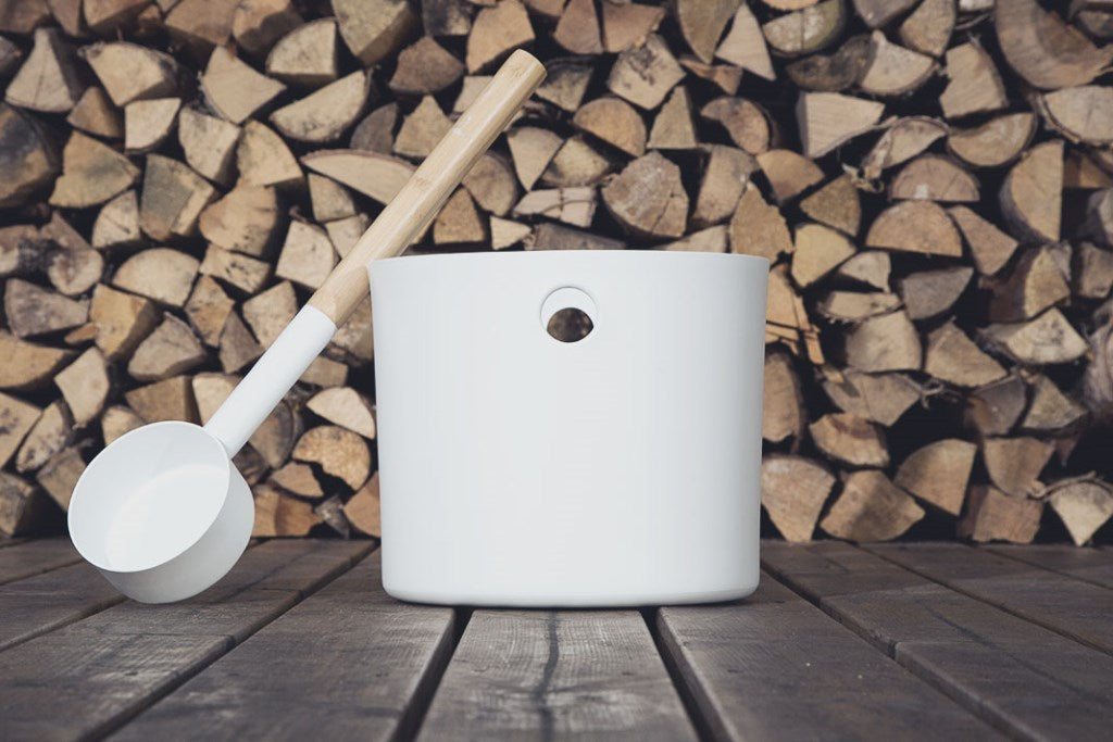 A stacked log background with a white bucket and ladle duo for the sauna sat on top of wooden slats