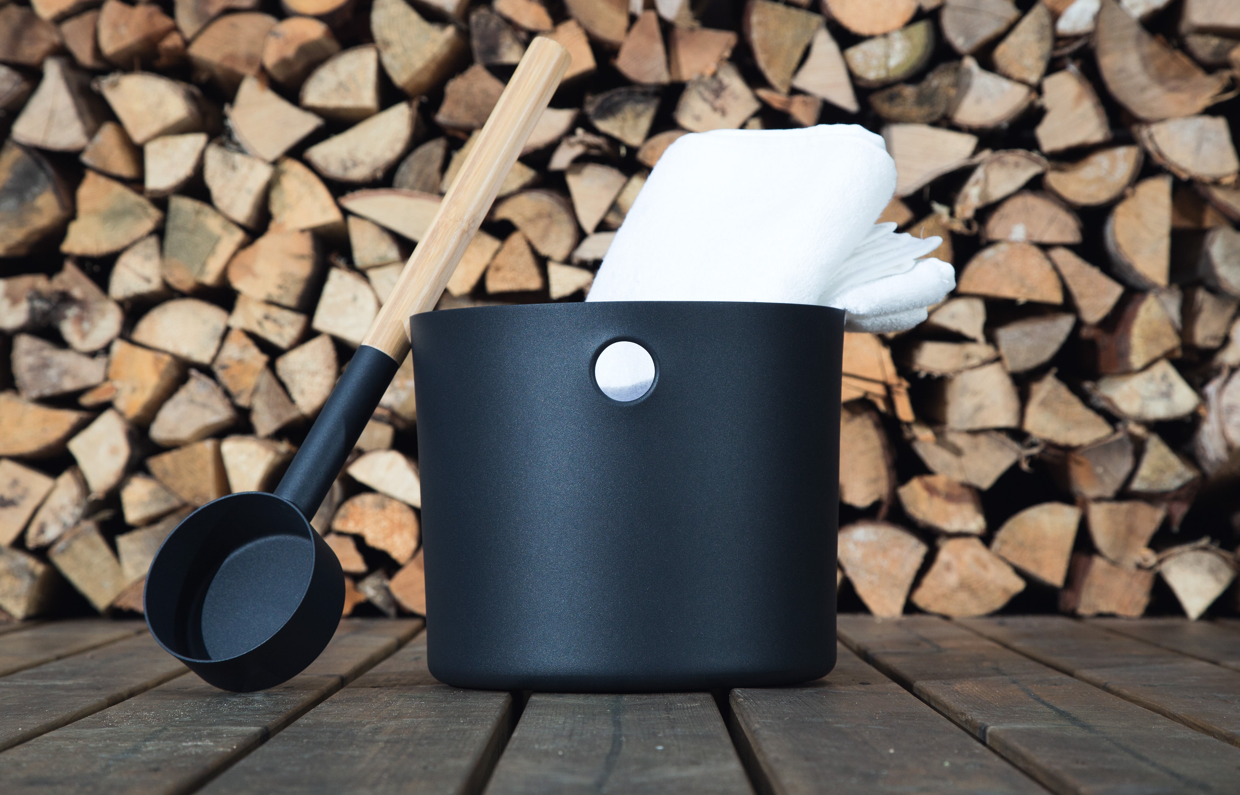 A stacked log background with a black bucket and ladle duo for the sauna sat on top of wooden slats