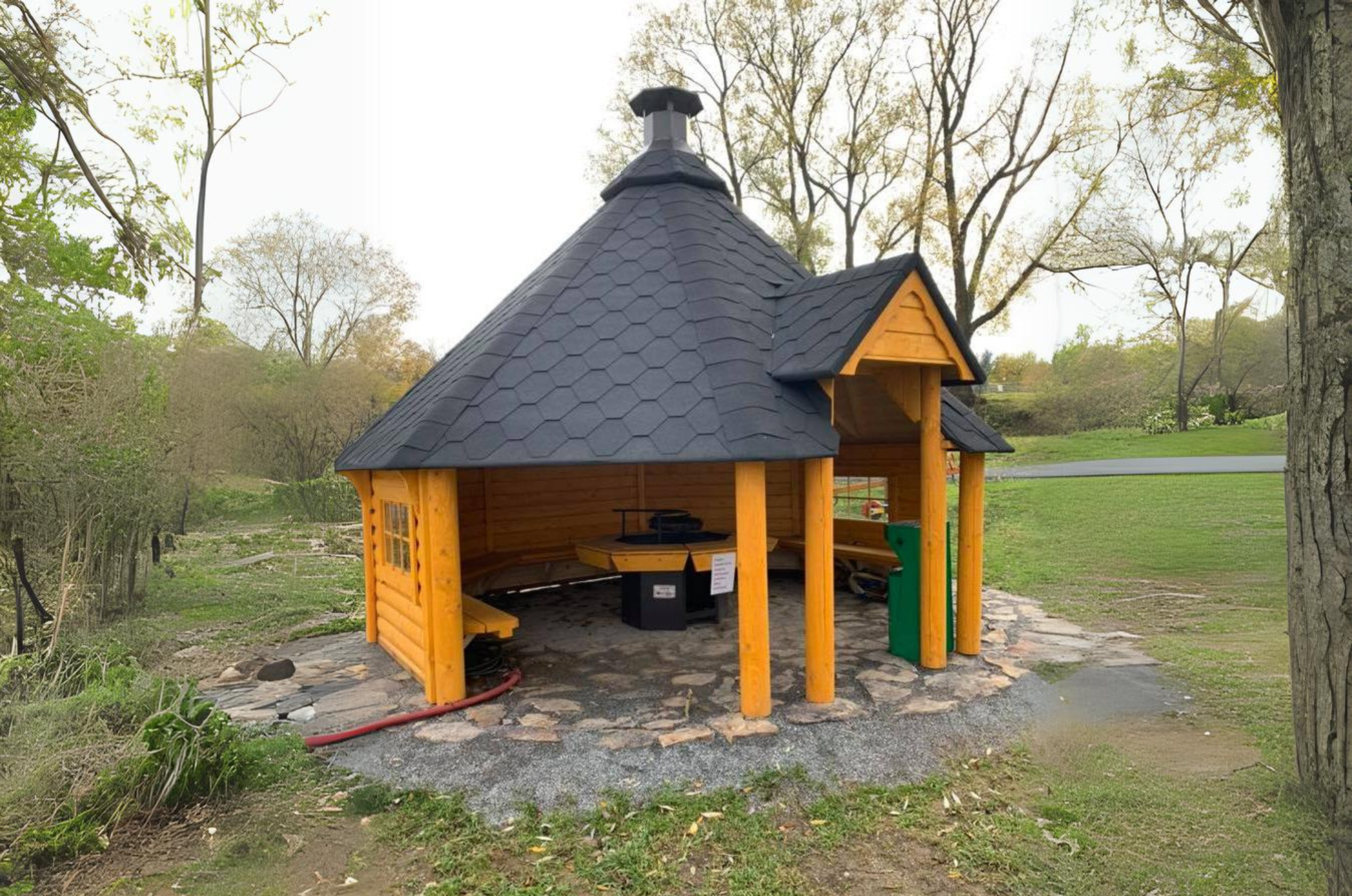 An Image showing the open BBQ hut with a black shingled roof in a garden space on a solid rock foundation with the Viking Industrier grill taking centre stage.