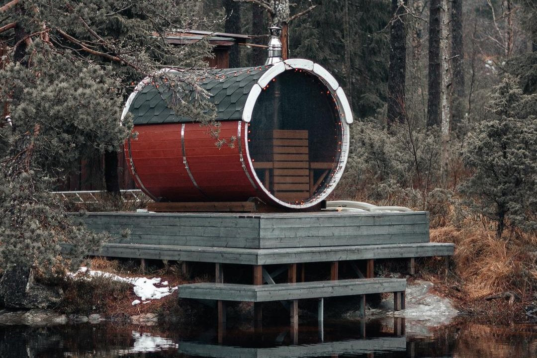 A red sided sauna barrel sat atop a wooden support overlooking a river trees and snow surround the sauna. 