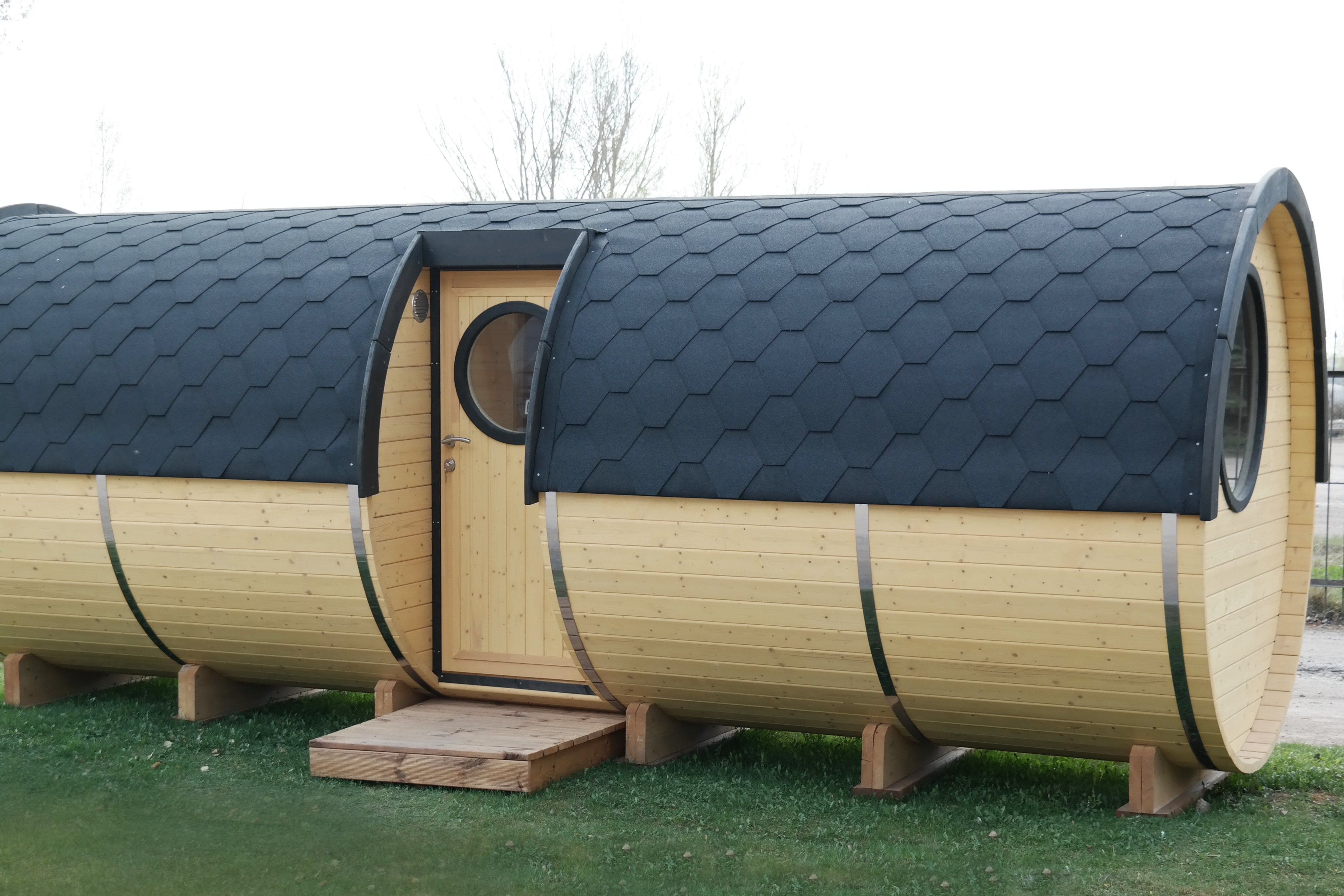 A side profile image showing the side entrance to the sauna barrel with a black shingled roof