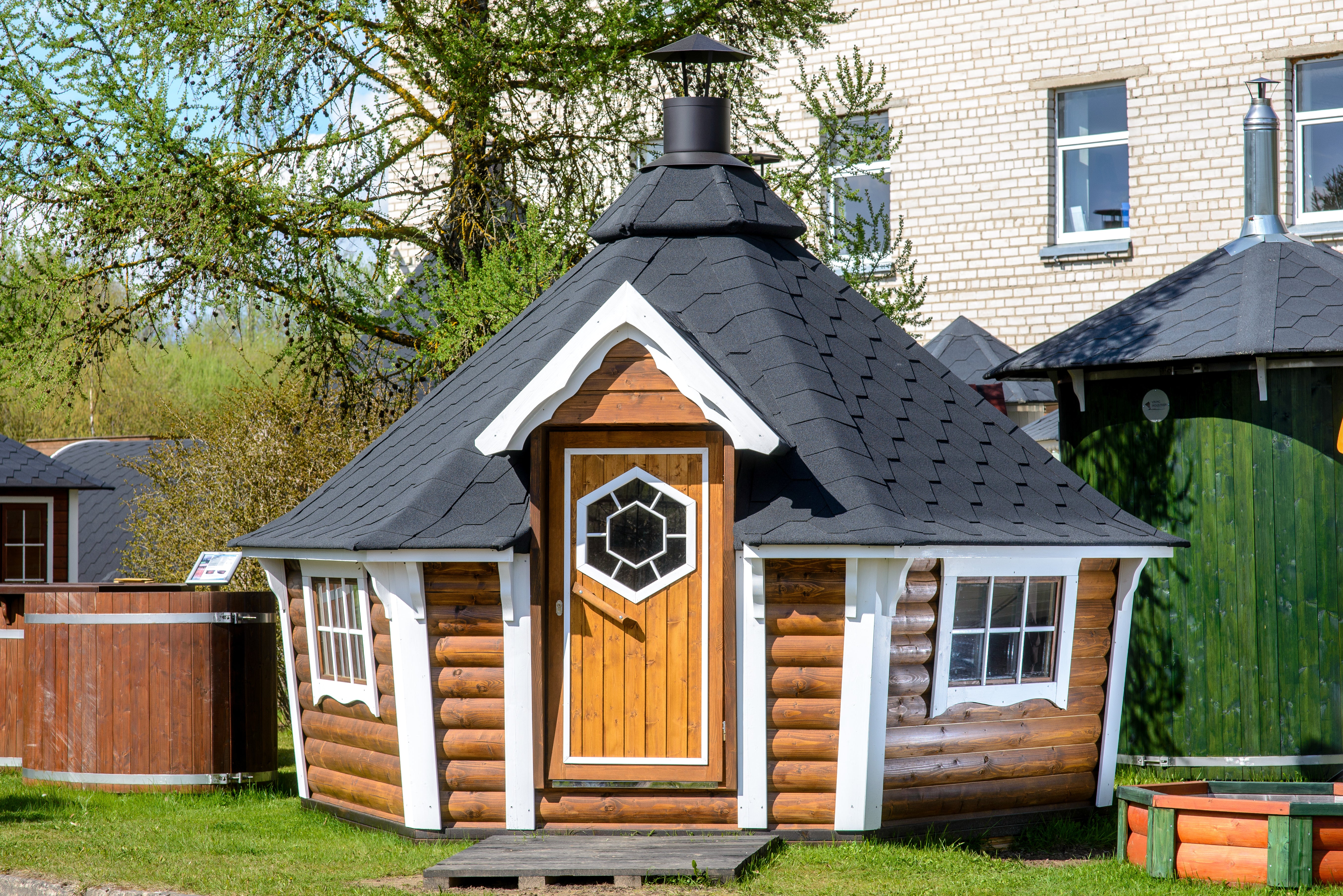 BBQ Hut with rustic oak (wood stain) walls and black shingle roof centred within a quaint garden, a hot tub to the left and a cabin to the right