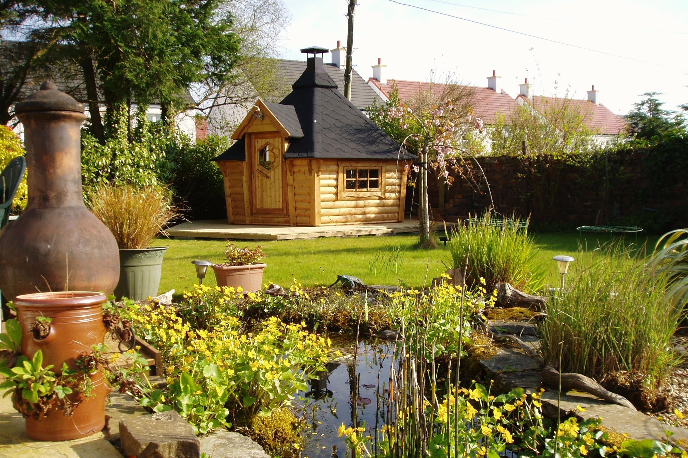 BBQ Hut with light oak (wood stain) walls and black shingle roof in the corner of a cottage garden featuring a pond and lots of plants