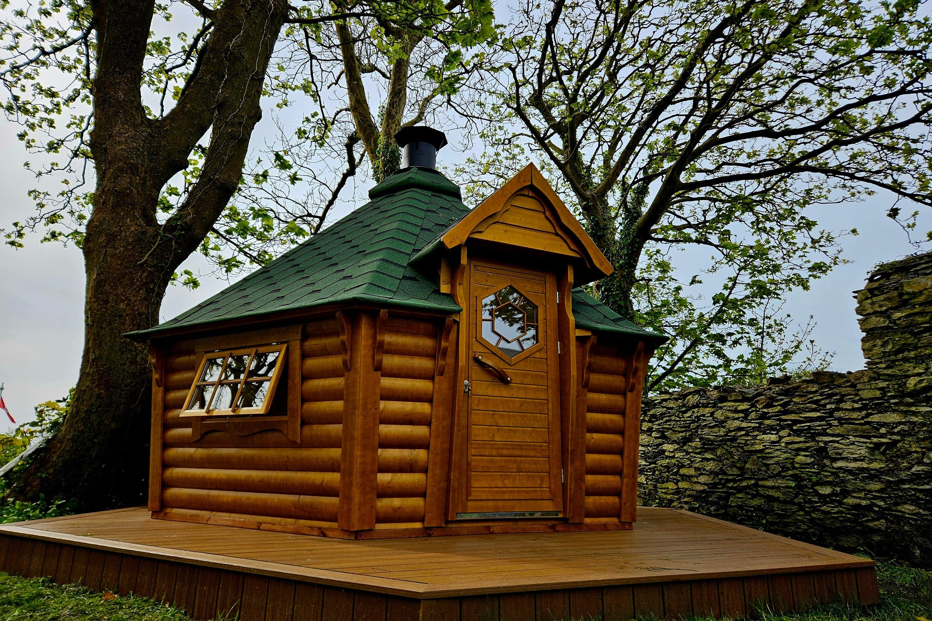 The BBQ Hut with rustic oak (wood stain) walls and green shingle roof atop a wooden terrace in a rainy garden